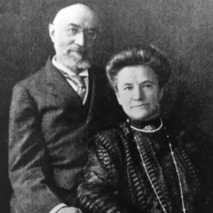 A posed studio photograph of two middle-aged people in the early 1900s. The man is seated behind the woman. He is light-skinned, bald, with a light beard and mustache, probably gray. He wears glasses and has a sharp, prominent nose. The expression on his face is pleasant as he looks into the camera. He wears a dark formal suit with a light shirt and dark necktie. Seated next to him is a light-skinned woman. Her dark hair is pulled back and arranged in Gibson Girl style. She has a pleasant look on her face, but she does not smile. Her hands are set in her lap. She wears a dark, high-necked, long-sleeved top and full skirt. There is a brooch at her neck, a strand of pearls around her neck, and a very long necklace that extends to her waist.