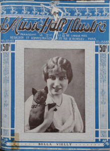 A young woman with light skin and short, dark hair holds a small dog. The woman is smiling at the camera. She wears a strand of pearls and a sheer top with elabororate decorations. The dog has dark fur and pricked ears. It is wearing a scarf with bells. The photo appears on the cover of a magazine about cabaret entertainment.