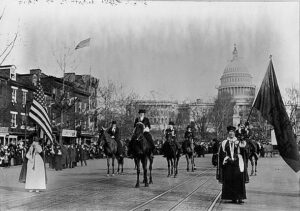 A parade of women on horseback and walking processes down Pennsylvania Avenue. The U.S. Capitol Building is in the distance. An American flag is seen in the foreground. 