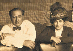 A man and woman sit in theater seats. The man has light skin and short dark hair. He wears a long-sleeved light shirt and a bow tie. The woman has light skin and dark hair. She wears a large hat with a feather and a long-sleeved top. They are posed with looking straight ahead with neutral expressions on their faces.