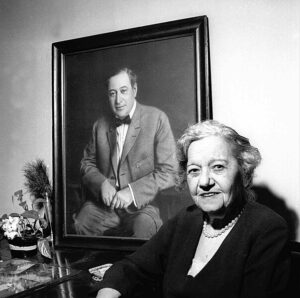 A posed snapshot of an older woman with short gray hair sits in front of a large framed photograph of her younger deceased husband, which is hanging on the wall. 