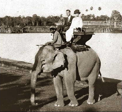 A woman and man ride atop an Asian elephant on a wooden bench-like saddle. A native guide also rides the elephant. The elephant is posed in front of a body of water. The surroundings are tropical, with palm trees in the distance.