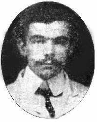 Studio photo of a man with light skin and dark hair. His hair is short and he has a full mustache. He is looking straight ahead with a neutral expression. He wears a light shirt and dark tie.