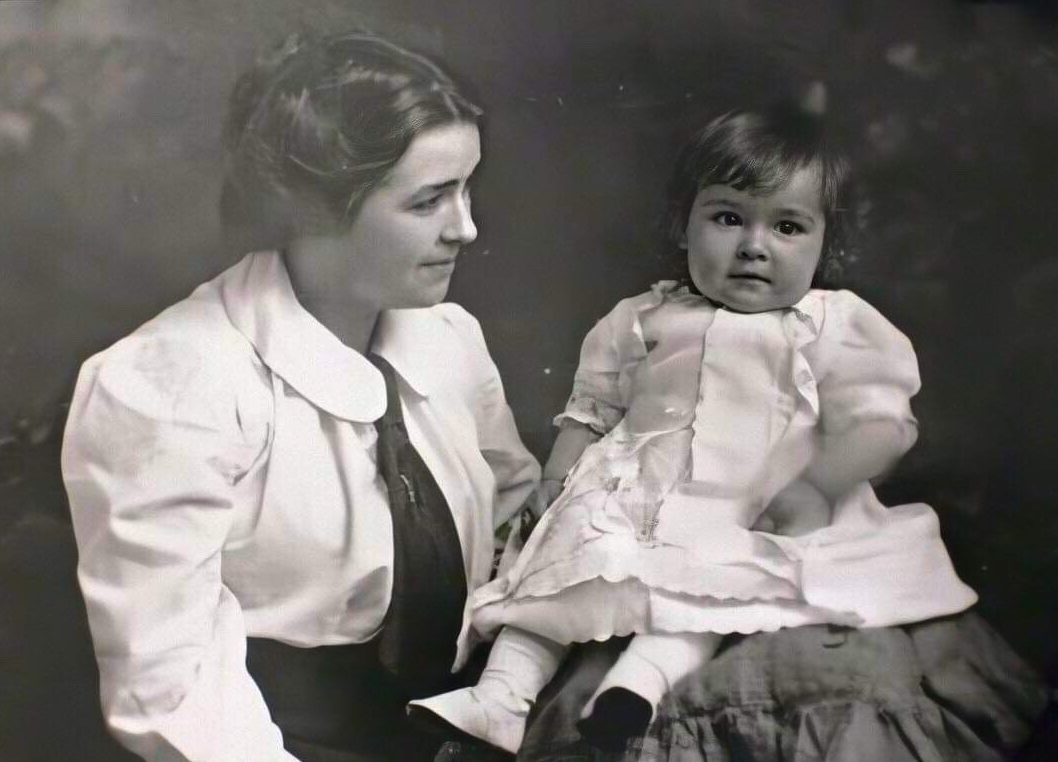 Studio photo of a young woman and toddler girl. The woman has long, dark hair pinned back in a bun. She has light skin. She wears a light long-sleeved shirt with large collar and a tie with a pin. The toddler is seated on a cushion and has light skin and short, dark hair. She wears a light-colored short-sleeved dress, light stockings, and light shoes.