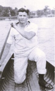 A young man with light skin and dark hair kneels in a rowboat. He wears a light short-sleeved shirt and light slacks, along with dark socks and dark dress shoes. He holds a boat paddle in his hands.